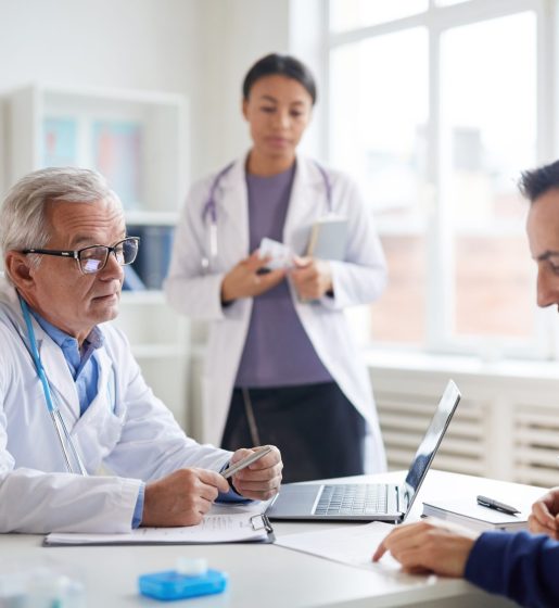 Senior male doctor in white coat talking to the patient at the table they discussing method of treatment with nurse in the background