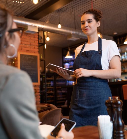 Young friendly waitress with touchpad standing by table with one of clients and taking her order on background of bar counter in restaurant